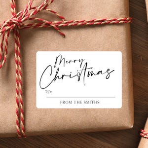 May include: A wrapped gift with a white rectangular label that reads "Merry Christmas" in elegant script. The label includes "TO:" and "FROM THE SMITHS" in a simple font. The gift is wrapped in brown paper and tied with red and brown twine.