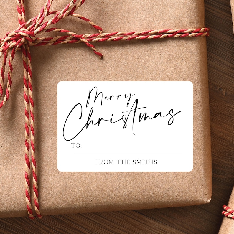 May include: A wrapped gift with a white rectangular label that reads "Merry Christmas" in elegant script. The label includes "TO:" and "FROM THE SMITHS" in a simple font. The gift is wrapped in brown paper and tied with red and brown twine.