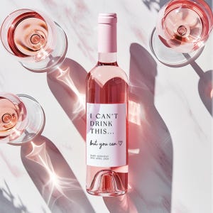 K&ouml;nnte beinhalten: Eine Glasflasche mit Ros&eacute;wein, einem hellrosa Verschluss und einem wei&szlig;en Etikett mit dem schwarzen Text I CAN'T DRINK THIS... but you can, einem Herzsymbol und dem Text BABY HANNIGAN DUE APRIL 2028, umgeben von drei Gl&auml;sern mit Ros&eacute;wein.