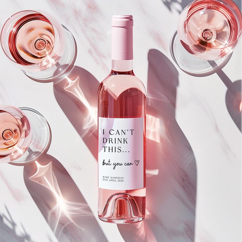 K&ouml;nnte beinhalten: Eine Glasflasche mit Ros&eacute;wein, einem hellrosa Verschluss und einem wei&szlig;en Etikett mit dem schwarzen Text I CAN'T DRINK THIS... but you can, einem Herzsymbol und dem Text BABY HANNIGAN DUE APRIL 2028, umgeben von drei Gl&auml;sern mit Ros&eacute;wein.