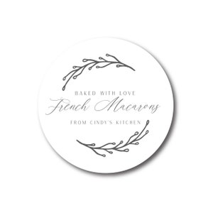 May include: A white, round sticker with the text "Baked with Love French Macarons From Cindy's Kitchen" in elegant script. The text is framed by a gray floral design. The sticker has a subtle shadow.