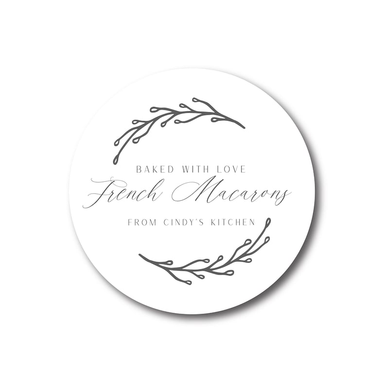 May include: A white, round sticker with the text "Baked with Love French Macarons From Cindy's Kitchen" in elegant script. The text is framed by a gray floral design. The sticker has a subtle shadow.