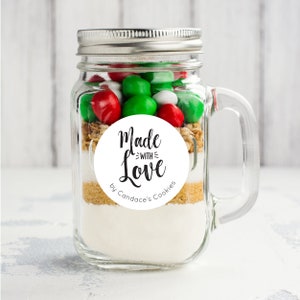 May include: A clear glass jar with a silver lid and a handle. The jar is filled with cookie ingredients, including white and red candy, brown sugar, and white flour. A white round sticker with black text reads "Made with Love by Candace's Cookies".