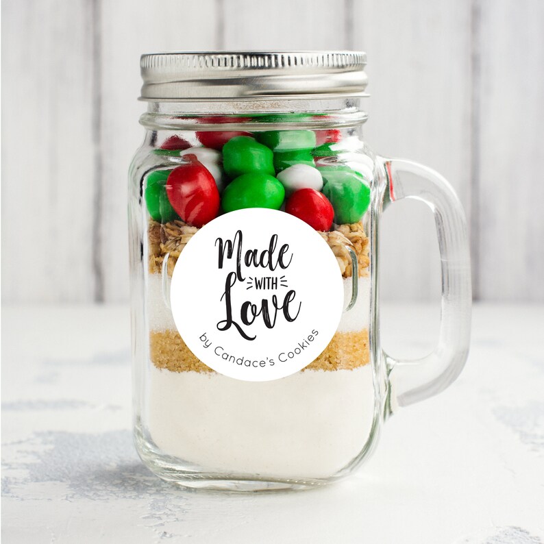 May include: A clear glass jar with a silver lid and a handle. The jar is filled with cookie ingredients, including white and red candy, brown sugar, and white flour. A white round sticker with black text reads "Made with Love by Candace's Cookies".