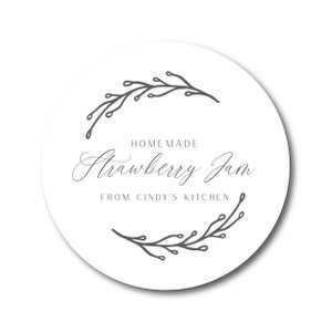 May include: A round white label with the text "Homemade Strawberry Jam From Cindy's Kitchen" in a script font. The label is encircled by a gray floral design.