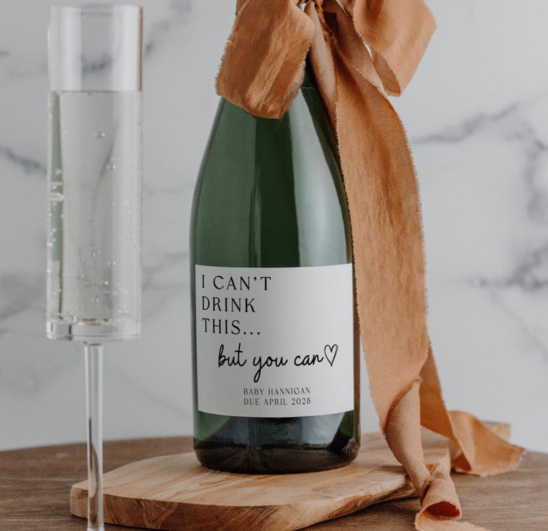 K&ouml;nnte beinhalten: Eine dunkelgr&uuml;ne Glasflasche mit einem wei&szlig;en Etikett, das den schwarzen Text I CAN'T DRINK THIS... but you can mit einem Herzsymbol und BABY HANNIGAN DUE APRIL 2026 tr&auml;gt. Ein langes, flie&szlig;endes kupferfarbenes Band ist um den Flaschenhals gebunden.