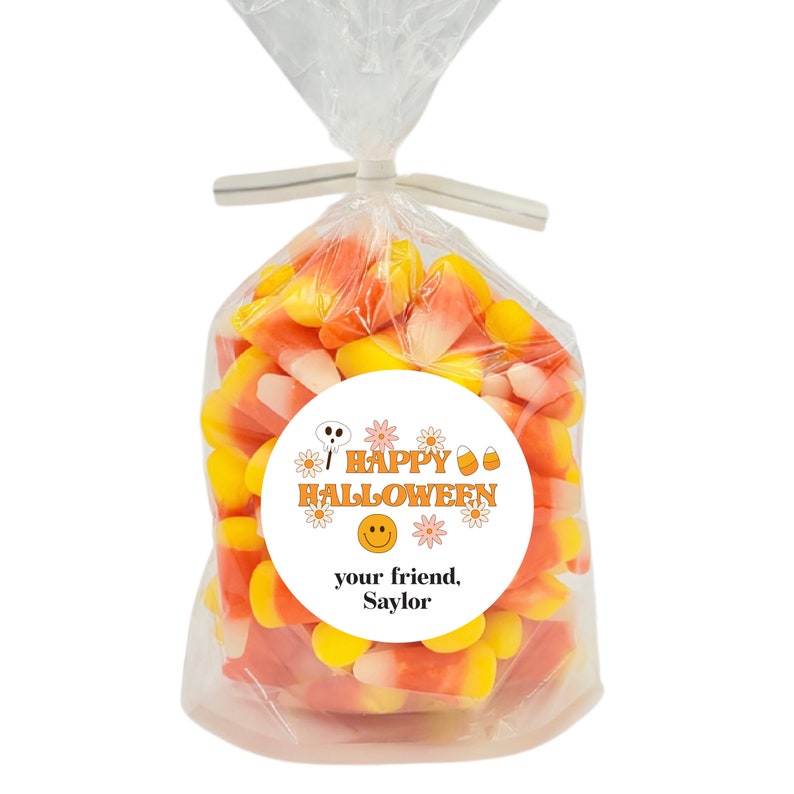 May include: A clear plastic bag filled with orange and yellow candy corn. A white round sticker with orange and pink flowers, a skull, a smiley face, and the text "Happy Halloween" and "your friend, Saylor" is attached to the bag.