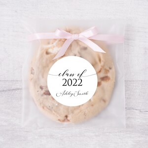May include: A white circle sticker with black text that reads "class of 2022" and "Ashley Smith". The sticker is on a clear plastic bag with a pink ribbon tied around it. The bag contains a cookie.