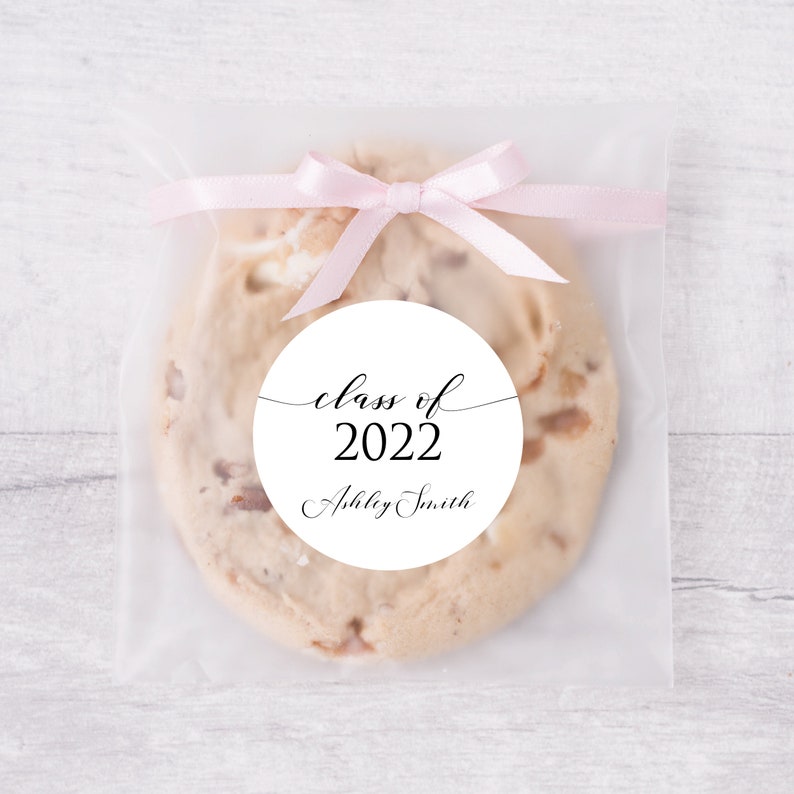 May include: A white circle sticker with black text that reads "class of 2022" and "Ashley Smith". The sticker is on a clear plastic bag with a pink ribbon tied around it. The bag contains a cookie.