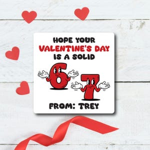 Pode incluir: Cart&atilde;o quadrado branco com o texto "HOPE YOUR VALENTINE'S DAY IS A SOLID 67 FROM: TREY" em vermelho e preto. Os n&uacute;meros 6 e 7 s&atilde;o personagens antropom&oacute;rficos. Decora&ccedil;&otilde;es em forma de cora&ccedil;&atilde;o vermelho e uma fita vermelha tamb&eacute;m est&atilde;o presentes.