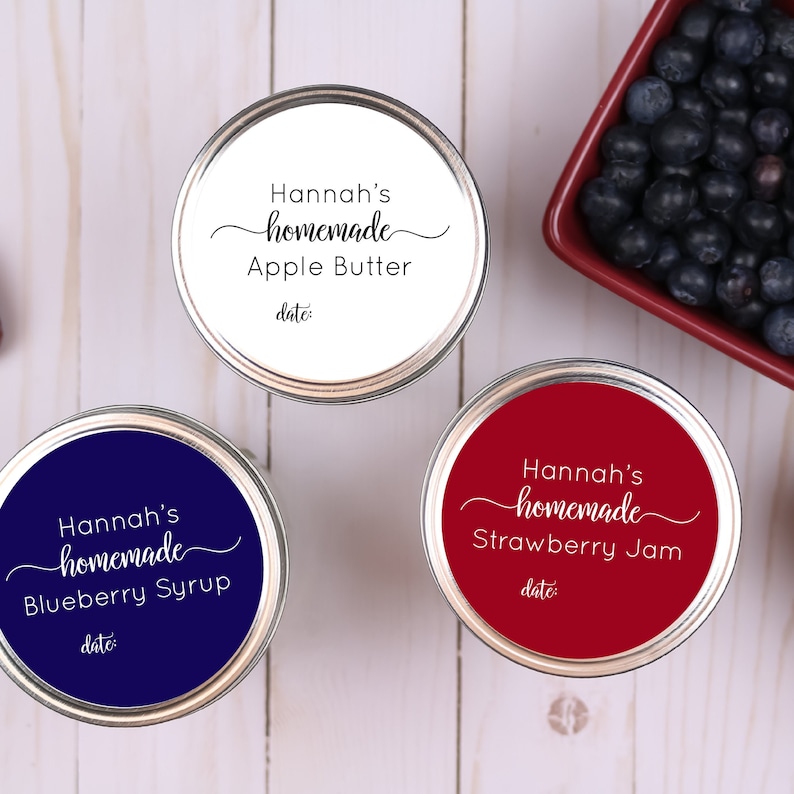 May include: Three jars of homemade preserves with white, red, and blue labels. The labels read "Hannah's homemade" with the names of the preserves: Apple Butter, Blueberry Syrup, and Strawberry Jam. A small dish of blueberries is in the background.