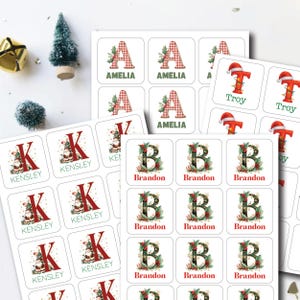 May include: A collection of personalized Christmas name stickers. The stickers feature letters with festive designs, including a Christmas tree, holly, and Santa hats. Names like Amelia, Kensley, Brandon, and Troy are displayed.