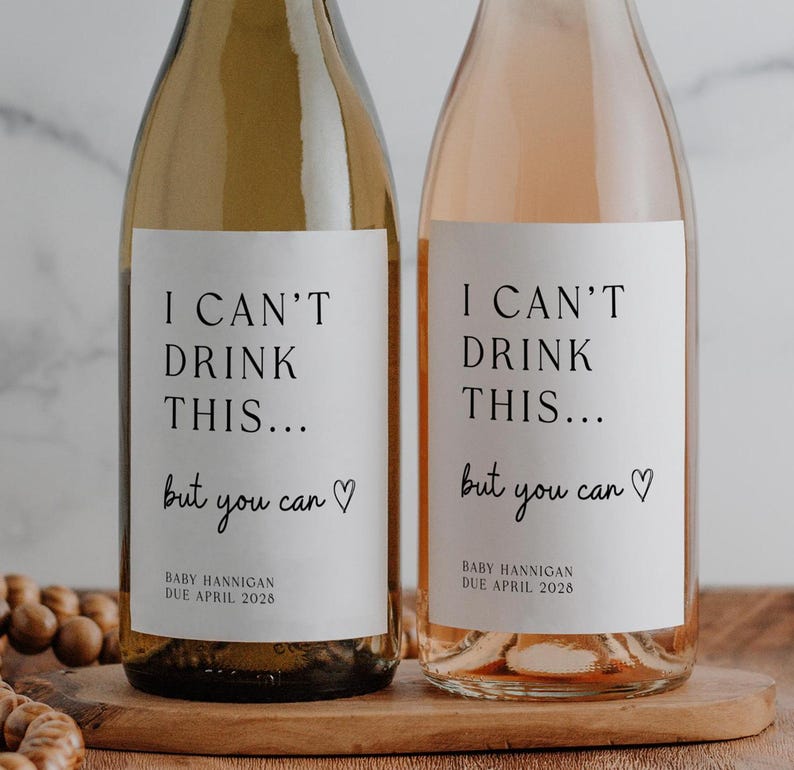 K&ouml;nnte beinhalten: Zwei Weinflaschen aus Glas, eine mit Wei&szlig;wein und eine mit Ros&eacute;wein, jeweils mit einem wei&szlig;en rechteckigen Etikett mit schwarzem Text I CAN'T DRINK THIS... but you can mit einem Herz und BABY HANNIGAN DUE APRIL 2028.