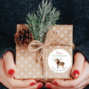 Op de afbeelding: Een bruine cadeaudoos met witte stippen is ingepakt met touw en een dennentak. Een ronde sticker met een cartoon-elch die een kerstmanmuts draagt en de tekst "Merry Christmasmoose From the Smith Family" is op de doos.