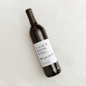 K&ouml;nnte beinhalten: Eine dunkle Glasweinflasche mit einem wei&szlig;en rechteckigen Etikett, das den schwarzen Text I CAN'T DRINK THIS... but you can, ein kleines Herzsymbol sowie den Text BABY HANNIGAN DUE APRIL 2028 in kleinerer Schriftgr&ouml;&szlig;e aufweist.