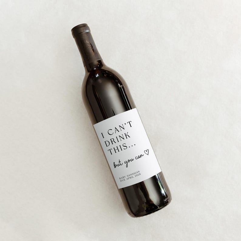 K&ouml;nnte beinhalten: Eine dunkle Glasweinflasche mit einem wei&szlig;en rechteckigen Etikett, das den schwarzen Text I CAN'T DRINK THIS... but you can, ein kleines Herzsymbol sowie den Text BABY HANNIGAN DUE APRIL 2028 in kleinerer Schriftgr&ouml;&szlig;e aufweist.