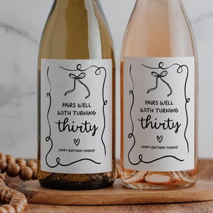 May include: Two glass wine bottles with white rectangular labels featuring a black hand-drawn border, a bow, and a heart. The text reads: PAIRS WELL WITH TURNING thirty, HAPPY BIRTHDAY NADINE! The bottles contain light and pink colored liquids.
