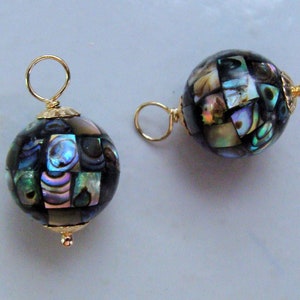 May include: Two abalone shell sphere pendants with gold-colored findings. The spheres are made of small, iridescent, square-shaped pieces of abalone shell, creating a mosaic effect. Each pendant has a loop at the top for hanging and a decorative gold-colored cap.