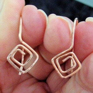 May include: A pair of handmade copper earrings. Each earring features a geometric, square spiral design. The earrings are held between fingers, showcasing their delicate, wire-like construction. The copper has a warm, reddish-brown hue.
