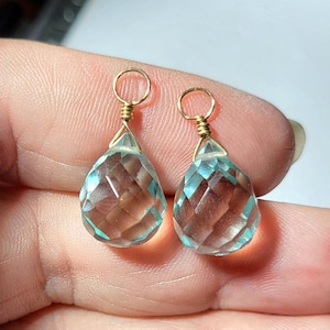 May include: Two faceted, pear-shaped light blue gemstone pendants, each topped with a gold-toned wire-wrapped loop. The translucent stones feature a geometric cut that reflects light, creating a clear, sparkling appearance in both pieces.