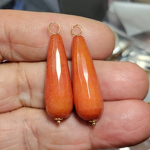 May include: Two faceted, teardrop-shaped orange stone pendants, each featuring a smooth, polished surface with subtle internal texture. Each pendant is topped with a small, circular gold-toned metal bail and finished with a tiny gold-toned bead at the base.
