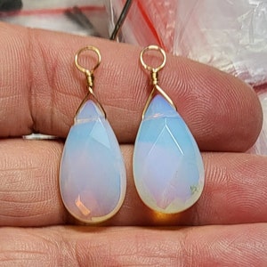 May include: Two pear-shaped, faceted opal-like pendants with a milky, translucent appearance and subtle blue and pink iridescence. Each pendant is topped with a gold-toned wire-wrapped bail featuring a small circular loop for attachment.