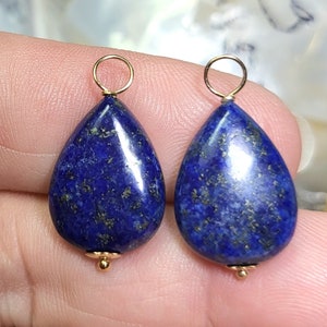 May include: Two teardrop-shaped lapis lazuli pendants with gold-colored findings. The deep blue stones have flecks of gold and are ready to be added to a necklace or earrings. The pendants are shown against a soft background.