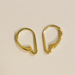 May include: Two gold-colored leverback earring findings. Each finding has a curved, D-shaped loop and a hinged lever closure. The findings are made of a shiny, metallic material and are set against a light background.