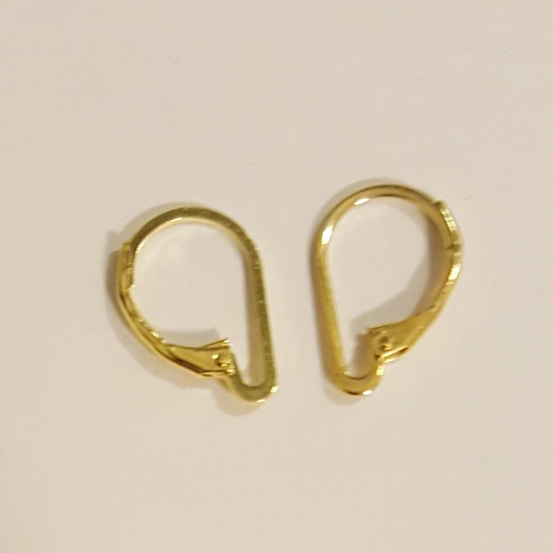 May include: Two gold-colored leverback earring findings. Each finding has a curved, D-shaped loop and a hinged lever closure. The findings are made of a shiny, metallic material and are set against a light background.
