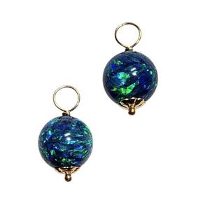 May include: A pair of spherical charms featuring a dark blue base with iridescent green and blue flecks. Each charm is topped with a gold-toned metal jump ring and finished with a small gold-toned metal cap at the bottom of the sphere.