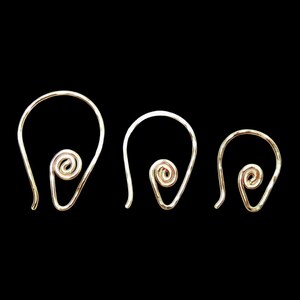 Puede incluir: Tres pendientes de alambre de color dorado con un diseño en espiral. Los pendientes están dispuestos de mayor a menor, cada uno con una parte superior curvada y una espiral en la parte inferior. El fondo es negro liso.