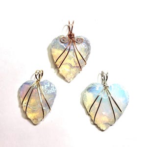 May include: Three heart-shaped pendants made of iridescent, translucent stone. Each pendant is wrapped in metal wire, with one in copper, one in gold-tone, and one in silver-tone wire. The stones exhibit a milky, opalescent sheen with subtle color shifts.