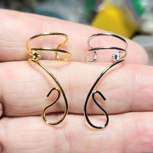 May include: Two ear cuffs, one gold-toned and one silver-toned, are displayed. Each cuff features a double-band design with a curved, decorative element. The gold cuff has a small, round accent, while the silver cuff has a small, clear gem.