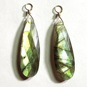 May include: Two teardrop-shaped labradorite pendants featuring faceted surfaces with iridescent green and gray flashes. Each gemstone is topped with a gold-toned wire-wrapped bail, creating a secure loop for attachment to jewelry chains or findings.
