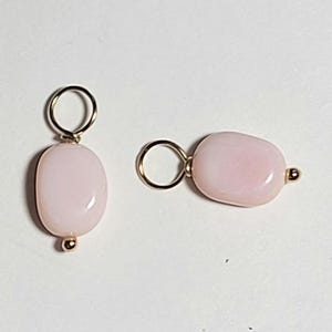 May include: Two pink oval-shaped gemstone charms with gold-colored metal loops and accents. The charms are smooth and polished, with a soft, light pink hue. The gold accents add a touch of elegance to the jewelry.