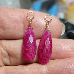 Może przedstawiać: Para kolczyków w kształcie łezki, o fasetowanej powierzchni, w żywym kolorze magenta. Kolczyki zawieszone są na złotych drucikach i kółeczkach. Kamienie szlachetne mają geometryczny szlif.