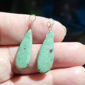 Peut inclure: Une paire de boucles d'oreilles en forme de goutte. Chaque boucle d'oreille présente une pierre vert clair avec une texture tachetée et une petite inclusion rouge foncé. Les pierres sont suspendues à des boucles en fil d'argent.