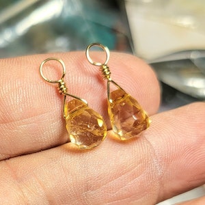 May include: A pair of gold-colored earrings featuring faceted, teardrop-shaped citrine gemstones. The citrine stones are suspended from gold-colored wire and small hoops. The earrings are displayed against a blurred background.