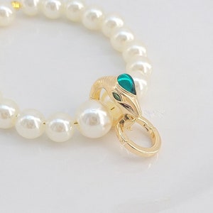 Könnte beinhalten: Ein Perlenarmband mit einem goldenen Schlangenverschluss. Der Schlangenkopf hat grüne und goldene Akzente. Das Armband besteht aus runden, weißen Perlen, die auf einer goldfarbenen Schnur aufgereiht sind.