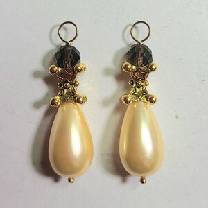 May include: A pair of elegant drop earrings. Each earring features a pearlescent, teardrop-shaped bead suspended below a gold-toned filigree design with a dark gray crystal and a gold hoop.
