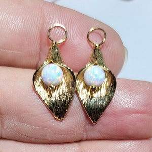 May include: A pair of gold-colored earrings featuring a calla lily design. Each earring has a textured, petal-shaped frame that cradles a round, iridescent opal-like bead. The earrings have a small loop at the top for attachment.