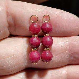 May include: A pair of dangle earrings featuring three faceted, deep red beads strung together with gold-colored accents. The earrings have a simple design with a small gold loop at the top for hanging.
