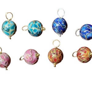 May include: A collection of eight colorful, round pendants with intricate enamel designs. The pendants feature a variety of colors, including turquoise, blue, pink, and red, with gold-colored metal accents and loops for hanging.