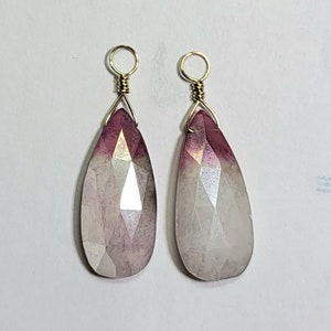 Puede incluir: Dos colgantes de gemas en forma de lágrima con superficies facetadas. Las gemas transicionan de un rosa intenso en la parte superior a un blanco translúcido en la parte inferior. Cada colgante está unido a un lazo de alambre dorado.