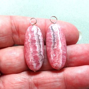 May include: Two oval-shaped rhodochrosite pendants featuring a polished surface with distinct bands of pink, white, and dark gray. Each stone is vertically oriented and topped with a small, circular silver-toned metal jump ring for attachment.