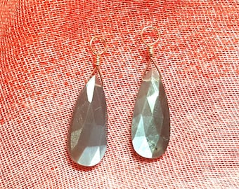 Può includere: Due pietre preziose grigie sfaccettate a forma di goccia, ciascuna sormontata da un occhiello in filo metallico color oro. Le pietre presentano una superficie liscia e lucida con molteplici sfaccettature geometriche che riflettono la luce, creando un riflesso sottile.
