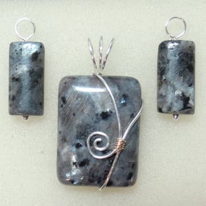 May include: A set of three labradorite pendants. The central pendant is rectangular with a silver wire wrap and a decorative swirl. Two smaller, cylindrical pendants with silver loops and silver beads complete the set.