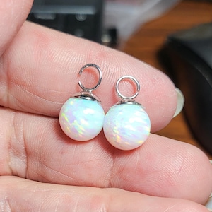 May include: Two spherical white opal charms featuring iridescent flashes of pink, blue, and green light. Each charm is topped with a small, circular silver-toned metal jump ring for attachment, showcasing a smooth, polished, and rounded surface.