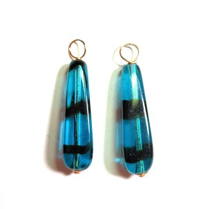 May include: A pair of drop-shaped pendants made of translucent turquoise glass, each featuring two horizontal black bands. The glass pieces are suspended from small, circular gold-toned metal jump rings attached to the top of each pendant.