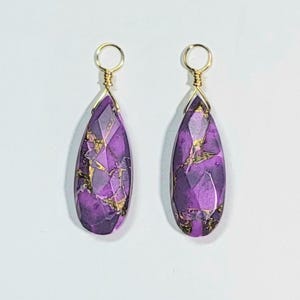 May include: Two teardrop-shaped pendants featuring faceted purple stone with metallic gold-colored veining. Each pendant is topped with a simple gold-colored wire loop bail for attachment, showcasing a polished finish and a distinct geometric pattern.